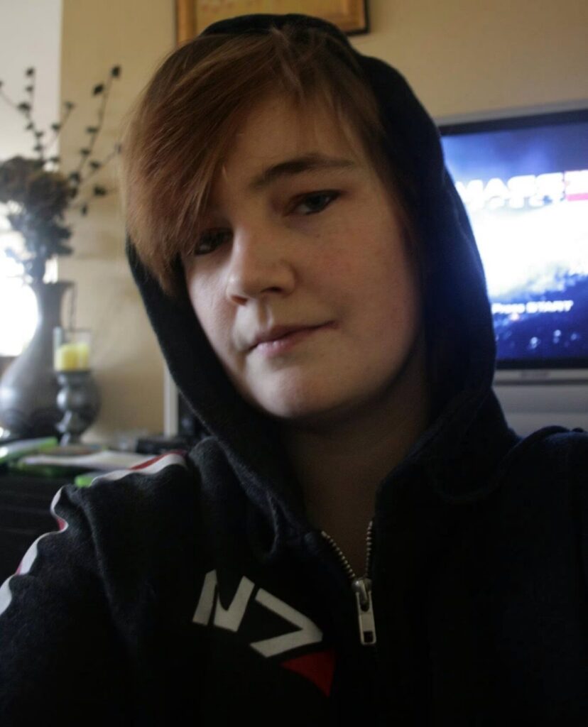 Foto van de auteur in Mass Effect N7-hoodie, met roodgeverfd haar en eyeliner. Op de achtergrond is het openingsscherm van Mass Effect 3 te zien.